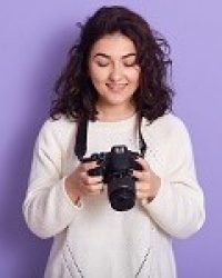 Professional photographer standing isolated over lilac background in studio, choosing photos, viewing works, smiling sincerely, being satisfied with result, having black curly hair. Skills concept.